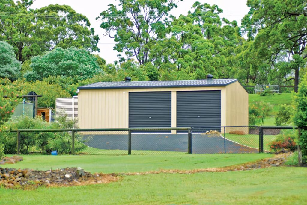 A beige double garage with black doors in a lush green garden.