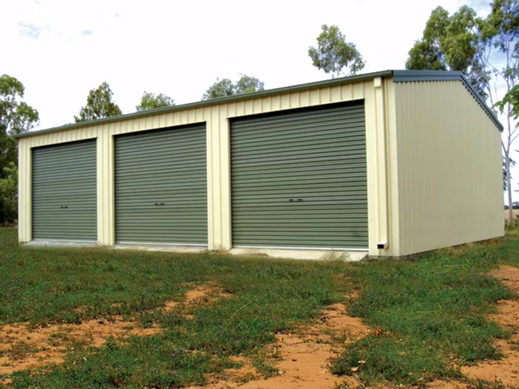 A large metal garage with three green roller doors in a grassy area.