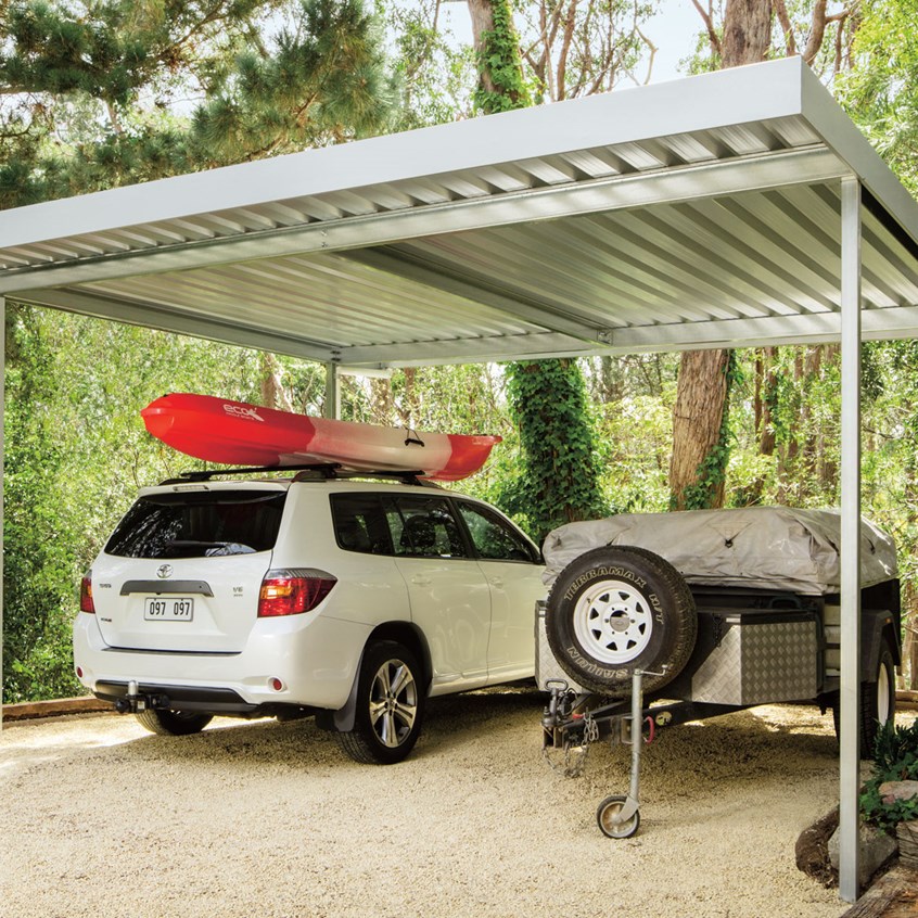 White SUV with red kayak on roof parked under metal carport beside covered trailer.